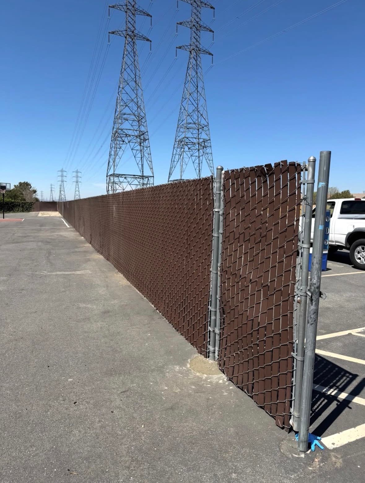 Brown chain-link fence with privacy slats along an asphalt lot under power line towers.