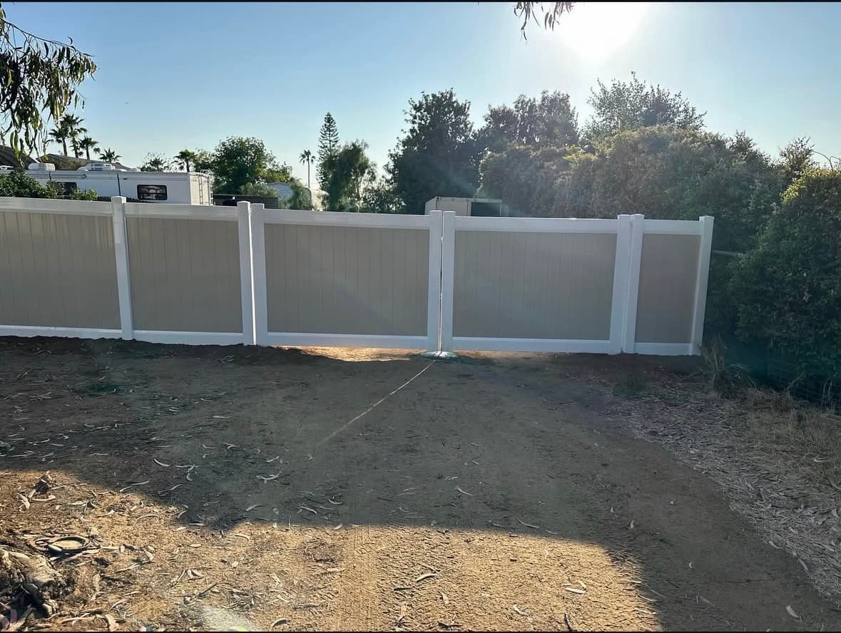 Tan and white vinyl double gate fence installed across a dirt driveway in bright sunlight.