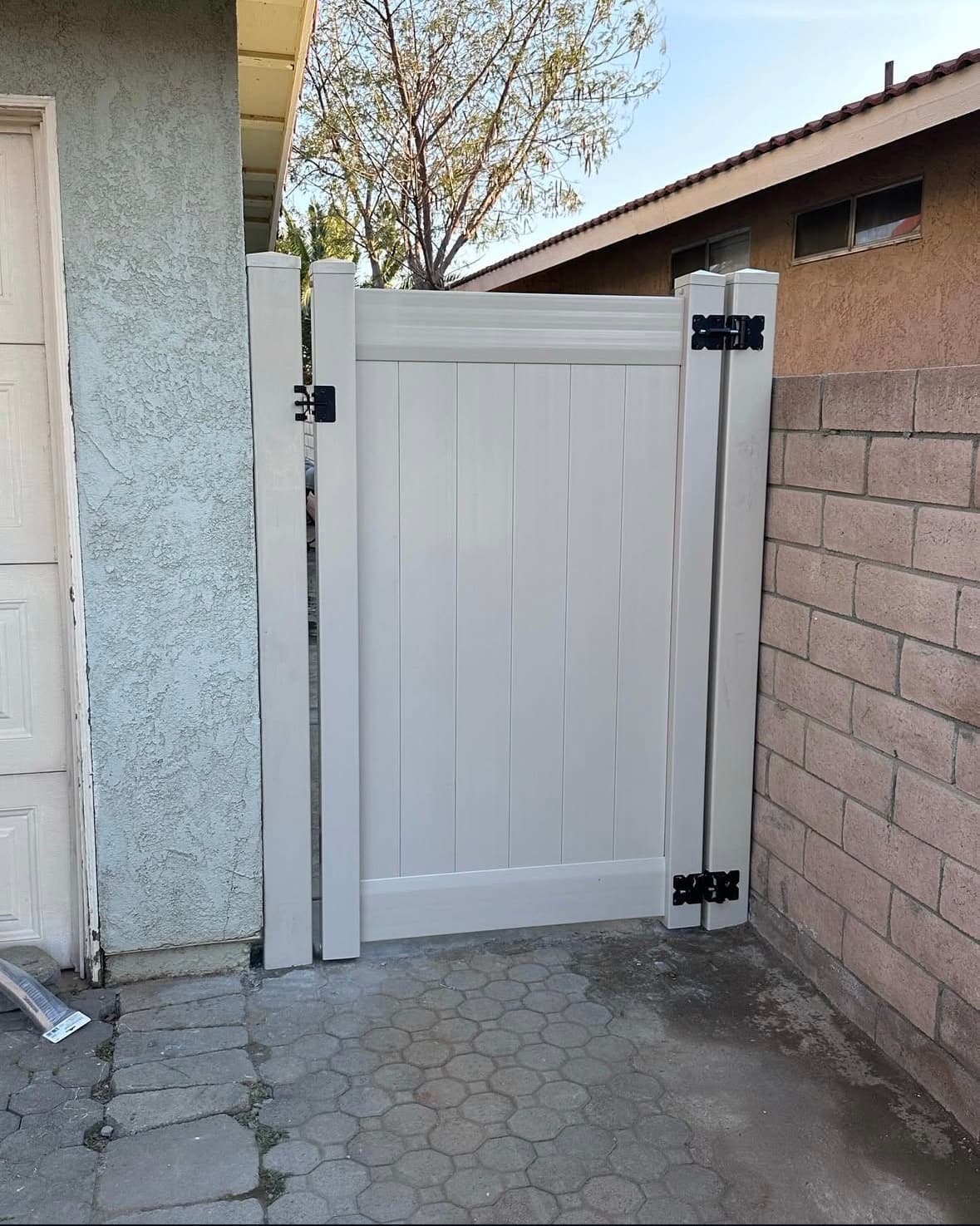 White vinyl privacy gate with black hinges between a stucco wall and brick fence.